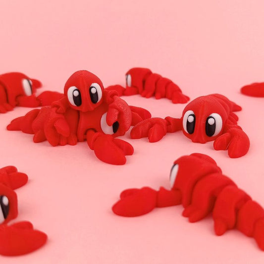 Several bright red 3D printed flexi lobsters with large cartoon-style eyes spread across a pink background, their claws and tails slightly curled to show off their flexible design.