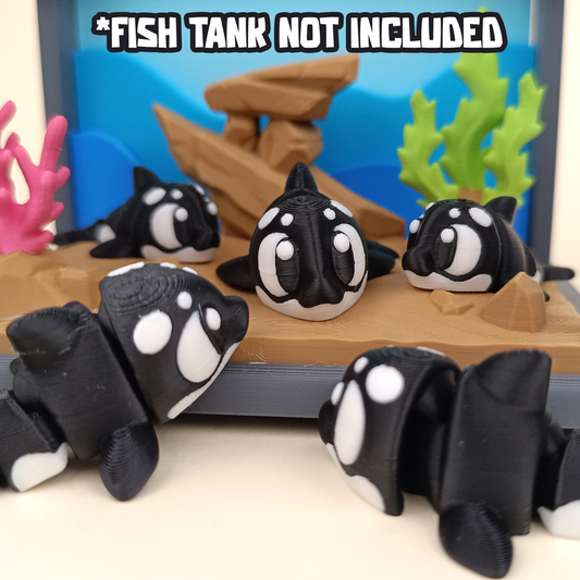 A group of small black-and-white 3D printed flexi orcas displayed inside a colorful aquarium-themed setup with coral decorations and a tan stand. Text above reads “Fish Tank Not Included.”