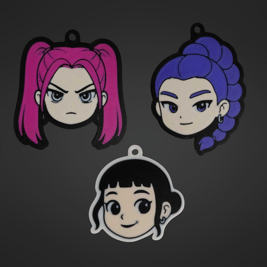 3D printed keychains of the K-Pop Demon Hunter trio – Mira with pink twin tails and a fierce expression, Rumi with long purple braided hair and a confident smile, and Zoey with short black hair and a cheerful expression. All three characters are displayed together on a dark background.