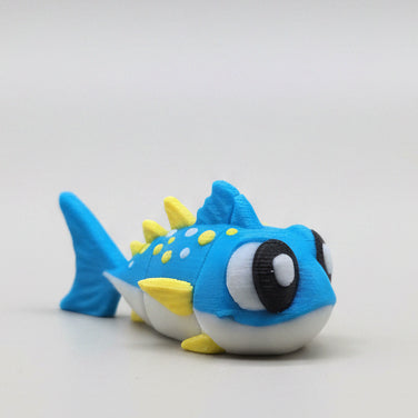 Cute 3D printed flexi tuna figure featuring bright blue colouring, yellow fin details, large expressive eyes, and an articulated flexible body. A fun ocean animal themed fidget toy and collectible photographed on a grey background.