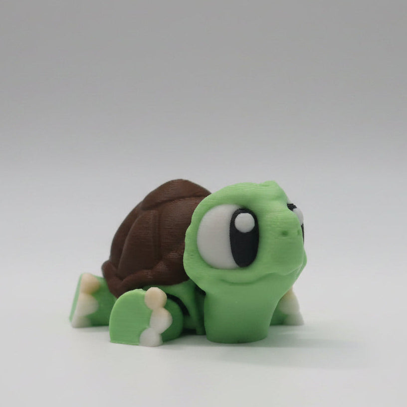 Green flexi 3D printed turtle figure shown from the side with large cartoon eyes, a brown detailed shell, and cream coloured feet. Cute articulated turtle collectible displayed on a simple white background.
