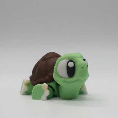 Green flexi 3D printed turtle figure shown from the side with large cartoon eyes, a brown detailed shell, and cream coloured feet. Cute articulated turtle collectible displayed on a simple white background.