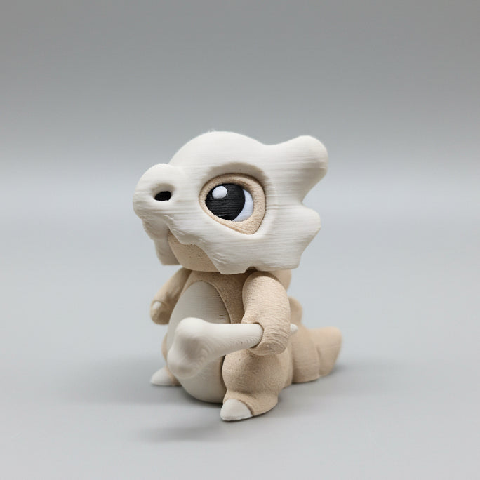 3D printed Cubone flexi figure in light beige tones, shown from a front-left angle holding a small bone, featuring large glossy eyes, a skull helmet, and articulated body joints on a neutral grey background