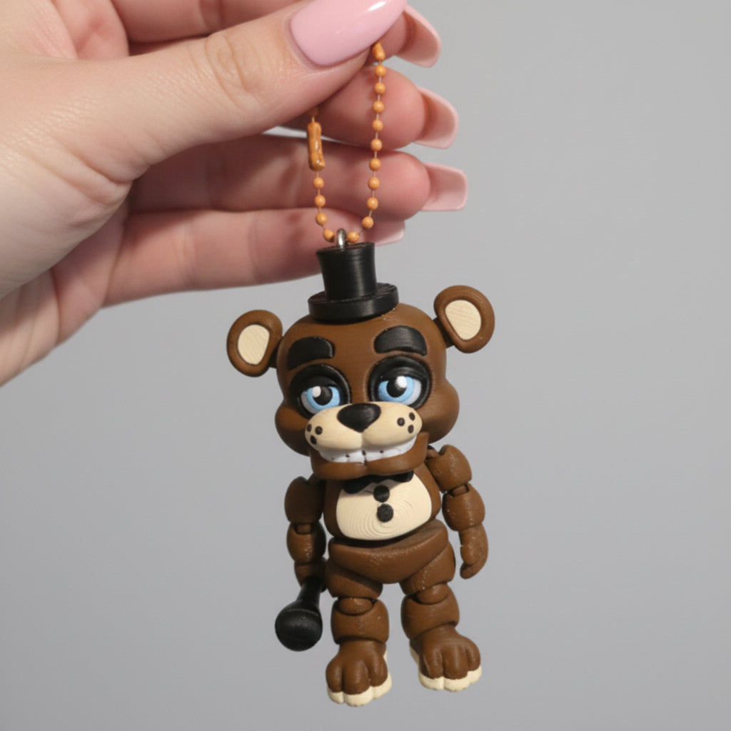 A hand holding a 3D printed Freddy Fazbear keychain inspired by Five Nights at Freddy’s. The brown animatronic bear features articulated limbs, a black top hat, bow tie, microphone, blue eyes, and button details, hanging from a gold ball chain against a light background.
