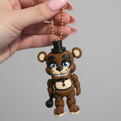 A hand holding a 3D printed Freddy Fazbear keychain inspired by Five Nights at Freddy’s. The brown animatronic bear features articulated limbs, a black top hat, bow tie, microphone, blue eyes, and button details, hanging from a gold ball chain against a light background.
