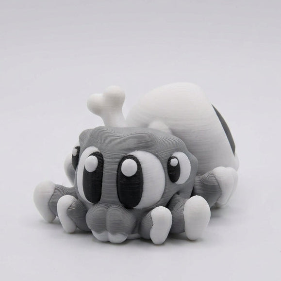 Side view of a cute grey 3D printed Spider Skull figurine showing large expressive eyes, small white-tipped legs, and a rounded white skull-shaped abdomen with a large black hollow eye socket, against a light grey background.
