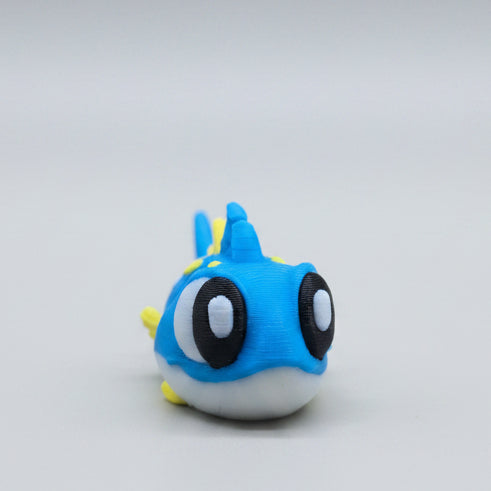 Front view of a cute 3D printed flexi tuna figure featuring bright blue colouring, yellow fins and spikes, big expressive eyes, and an adorable ocean-inspired design. A flexible articulated sea animal fidget toy and collectible photographed on a grey background.