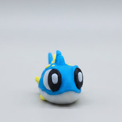 Front view of a cute 3D printed flexi tuna figure featuring bright blue colouring, yellow fins and spikes, big expressive eyes, and an adorable ocean-inspired design. A flexible articulated sea animal fidget toy and collectible photographed on a grey background.
