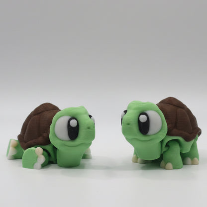 Two green flexi 3D printed turtle figures facing each other with large cartoon eyes, brown textured shells, and cream coloured feet. Cute articulated turtle collectibles displayed together on a plain white background.