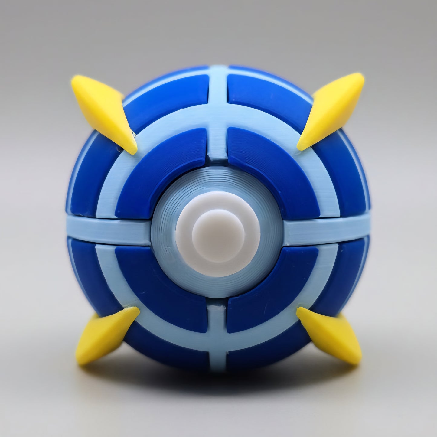 Front view of a 3D printed Beast Ball featuring dark blue and light blue segmented panels, white centre button, and four yellow spike accents on a clean white background.